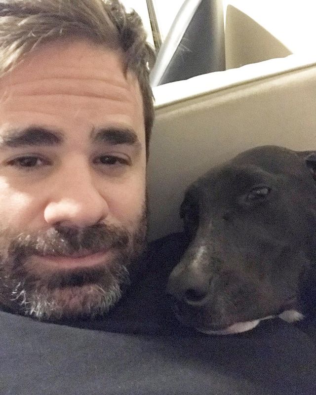 Yannis Pappas in a black t-shirt posing with his pet dog.